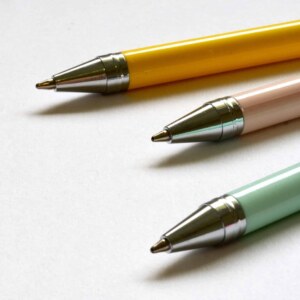 Three multicolored ballpoint pens lay on a clean white surface in a minimal style.